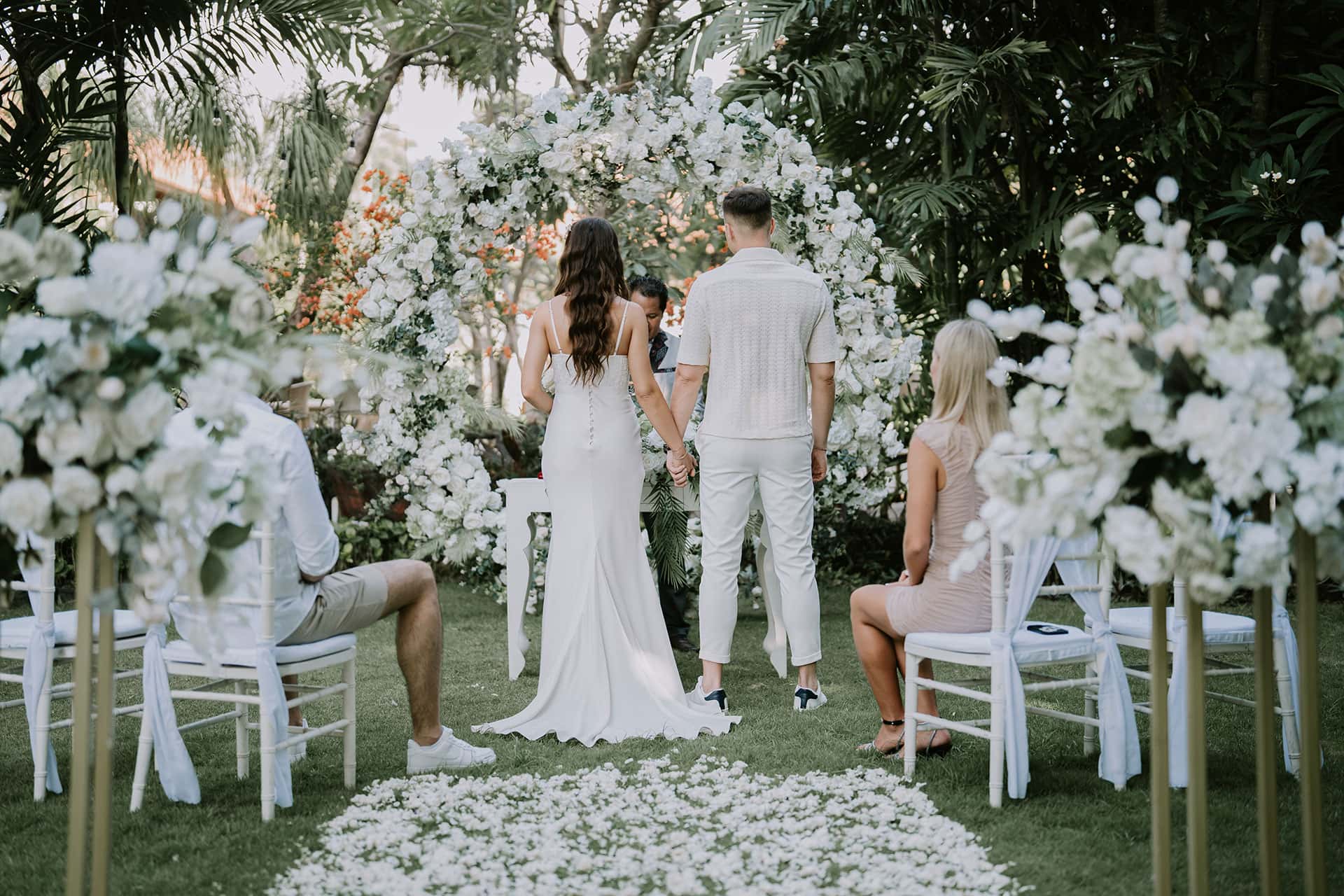Elegant outdoor wedding ceremony in white featuring a large floral arch and petal aisle.