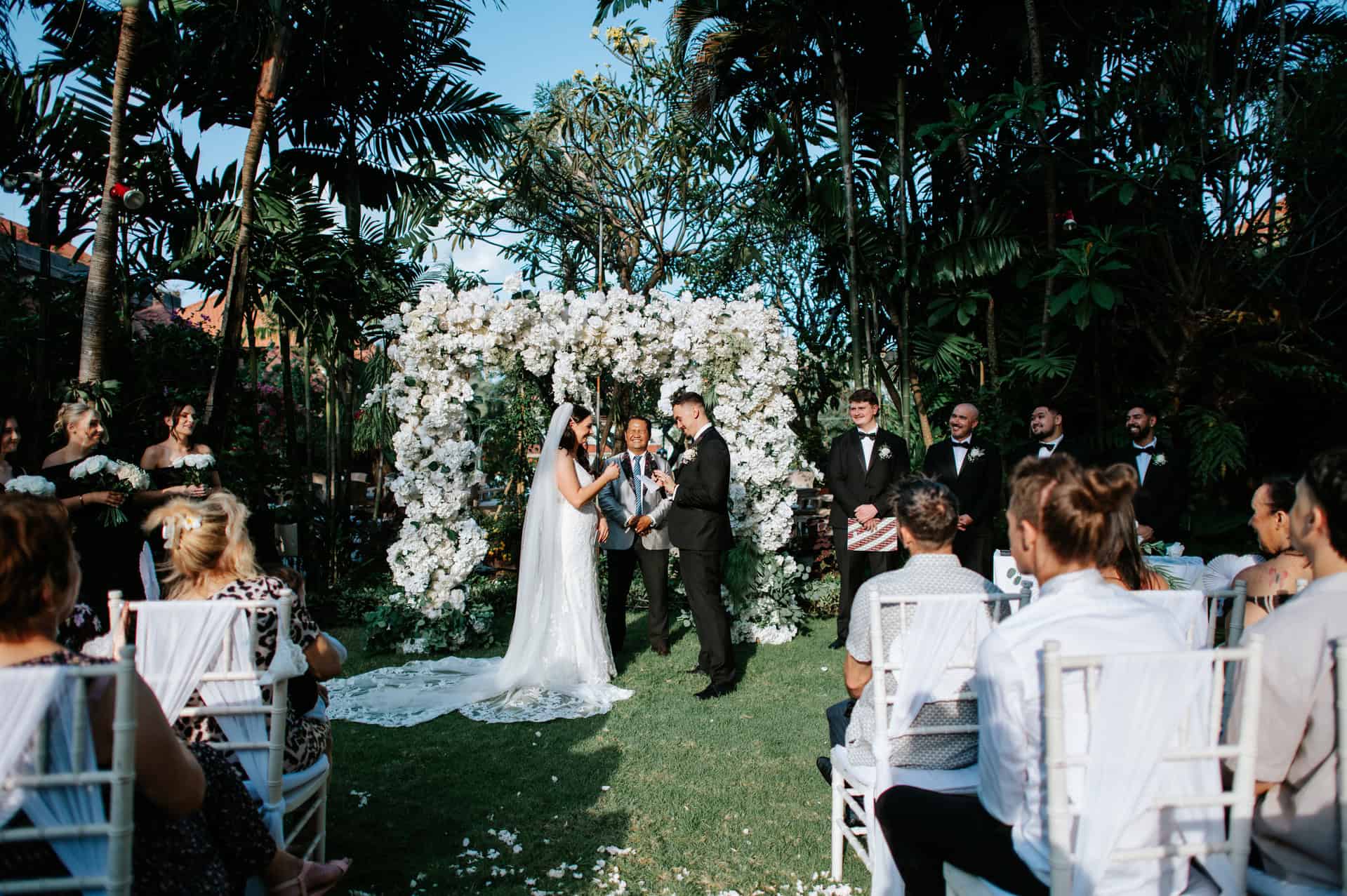 Garden holy matrimony ceremony at Banyan Garden with white floral arch altar, Tiffany chairs seating, and tropical greenery setting for Bali garden wedding venue in Legian