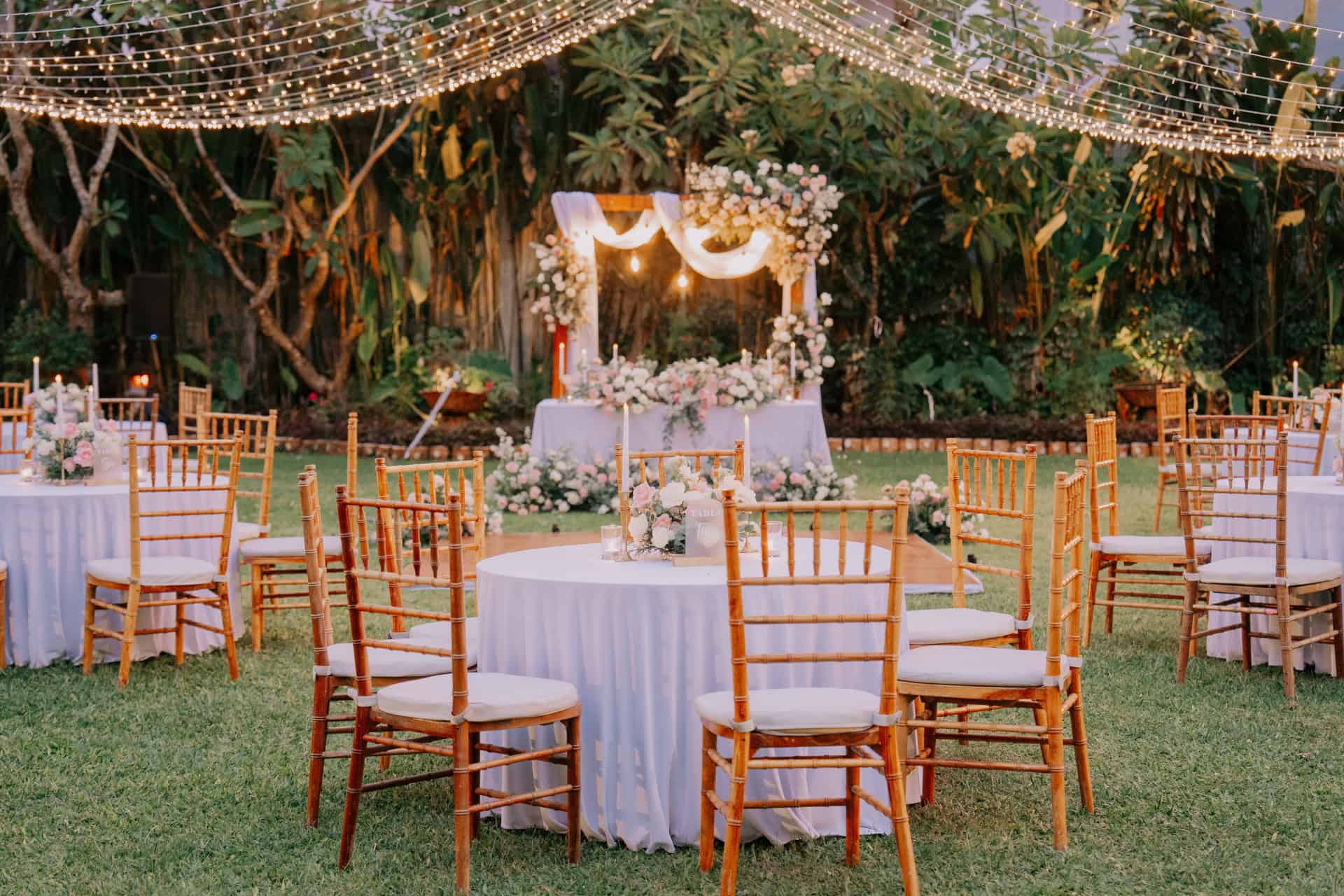 Elegant garden wedding reception setup at Astagina Courtyard with round tables, gold Chiavari chairs, fairy light canopy, and floral altar backdrop at a Bali garden wedding venue in Legian